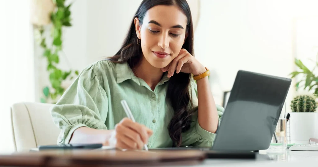 Erwachsene sitzt an einem Tisch, schreibt mit einem Stift auf ein Notizbuch, vor ihr steht ein aufgeklappter Laptop, im Hintergrund Pflanzen und ein Kaktus auf dem Tisch