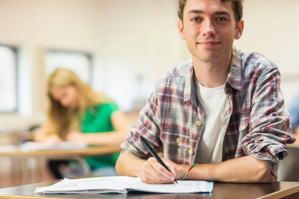 Junger Mann in kariertem Hemd schreibt in einem Klassenzimmer in ein Heft