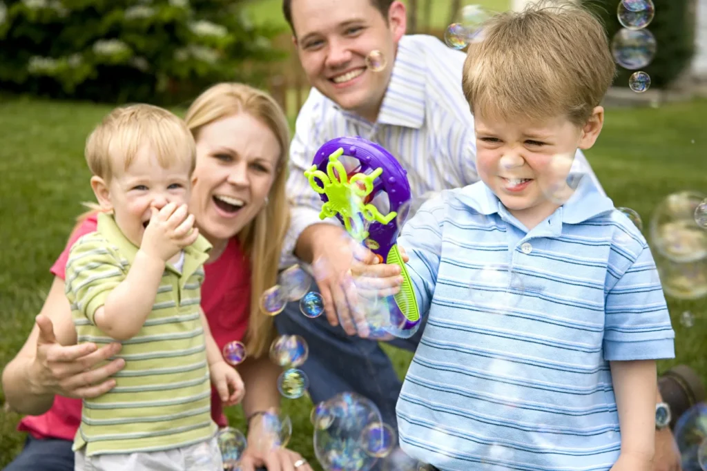 Zwei Kinder und zwei Erwachsene im Hintergrund spielen mit einer Seifenblasenpistole im Freien